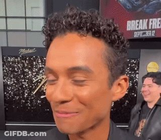 A Man With Curly Hair Is Smiling With His Eyes Closed In Front Of A Sign That Says Break Free GIF