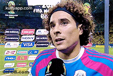 A Man With Curly Hair Is Talking Into A Microphone On A Soccer Field GIF