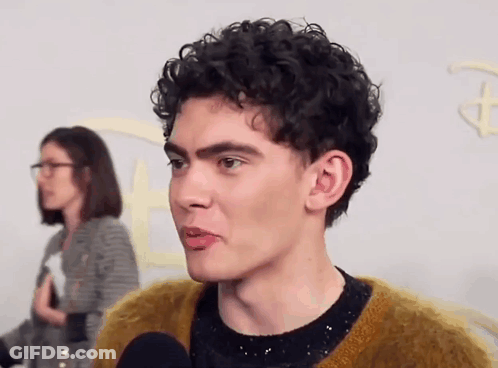 A Man With Curly Hair Is Talking Into A Microphone While A Woman Stands Behind Him . GIF