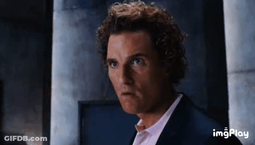 A Man With Curly Hair Is Wearing A Suit And A Pink Shirt And Making A Funny Face . GIF