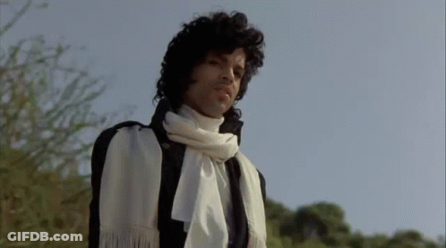 A Man With Curly Hair Is Wearing A White Scarf Around His Neck And A Black Jacket . GIF