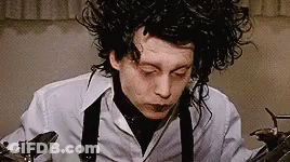 A Man With Curly Hair Is Wearing A White Shirt And Suspenders And Holding A Pair Of Scissors . GIF