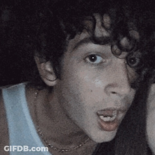 A Man With Curly Hair Is Wearing A White Tank Top And A Necklace GIF