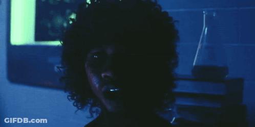 A Man With Curly Hair Looks At The Camera In A Dark Room GIF
