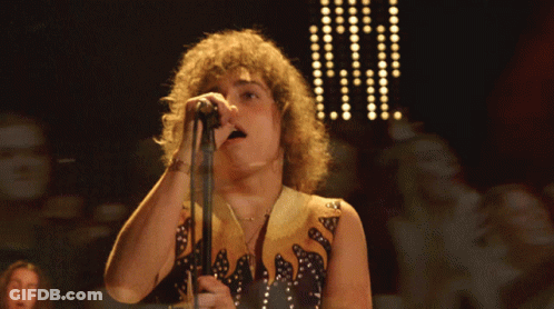 A Man With Curly Hair Singing Into A Microphone While Wearing A Black And Gold Outfit GIF
