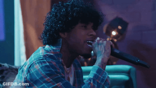 A Man With Curly Hair Singing Into A Microphone While Wearing A Plaid Shirt GIF