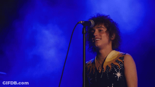 A Man With Curly Hair Singing Into A Microphone With A Blue Background GIF