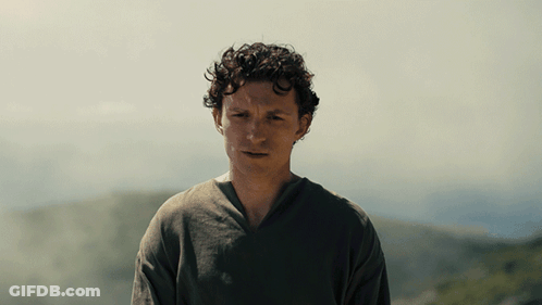 A Man With Curly Hair Wearing A Grey Shirt Stands In Front Of A Mountain GIF