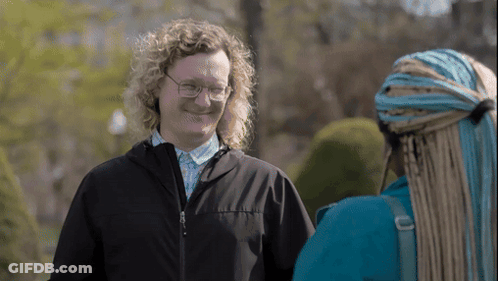 A Man With Curly Hair Wearing Glasses And A Black Jacket Talks To A Woman With Braids GIF
