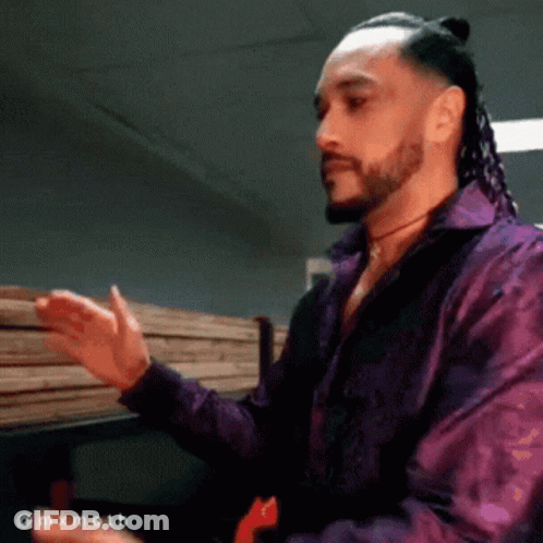 A Man With Dreadlocks And A Beard Wearing A Purple Shirt Is Standing In Front Of A Stack Of Wood GIF