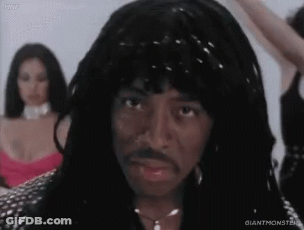 A Man With Dreadlocks And A Mustache Is Standing In Front Of Two Women In Dresses . GIF