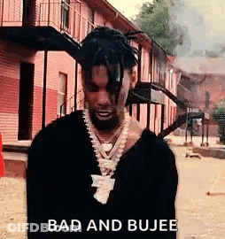 A Man With Dreadlocks And A Necklace Is Standing In Front Of A Building Smoking A Cigarette . GIF