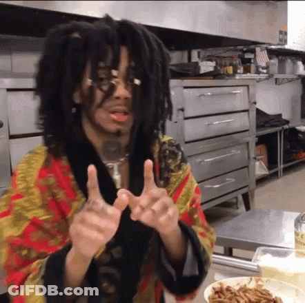 A Man With Dreadlocks And Glasses Is Giving Two Thumbs Up In A Kitchen . GIF