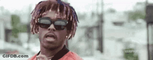 A Man With Dreadlocks And Sunglasses Is Standing In Front Of A Building And Looking At The Camera . GIF