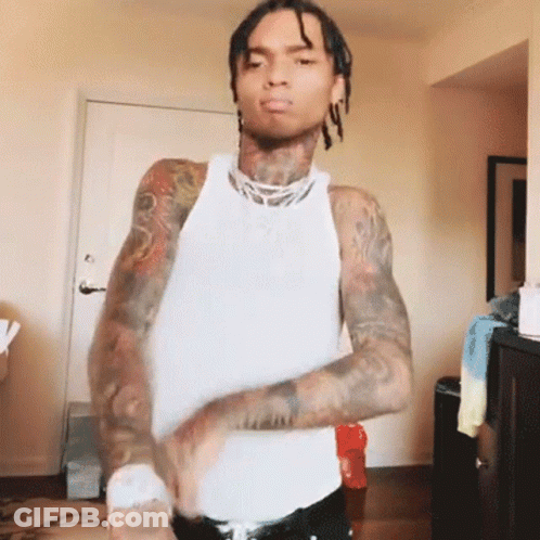 A Man With Dreadlocks And Tattoos Is Wearing A White Tank Top And Black Pants . GIF