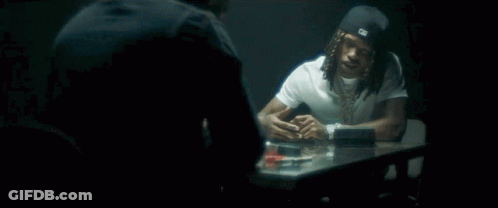 A Man With Dreadlocks Is Sitting At A Table Talking To Another Man In A Dark Room . GIF