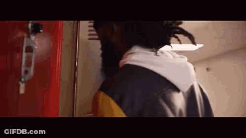 A Man With Dreadlocks Is Standing In Front Of A Red Locker In A Hallway . GIF
