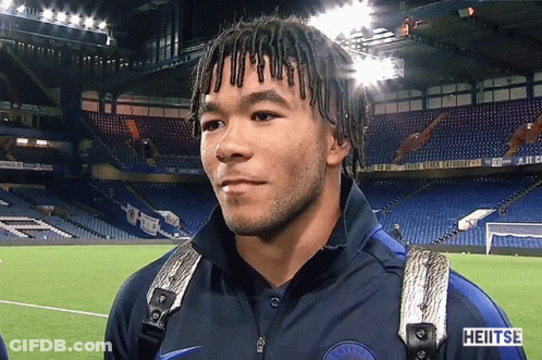 A Man With Dreadlocks Is Standing In Front Of A Stadium That Says Heitse GIF