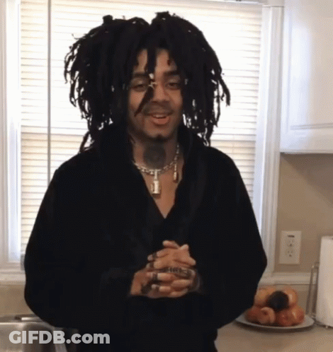 A Man With Dreadlocks Is Standing In Front Of A Window Holding A Glass GIF