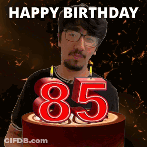 A Man With Glasses And A Beard Is Holding A Birthday Cake With Candles In The Shape Of The Number 85 GIF