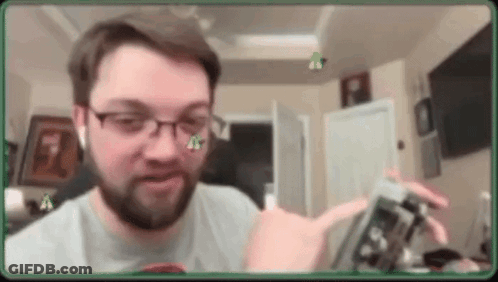 A Man With Glasses And A Beard Is Holding A Cell Phone In A Room . GIF