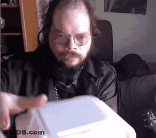 A Man With Glasses And A Beard Is Holding A Styrofoam Box GIF