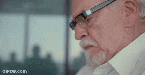 A Man With Glasses And A Beard Is Looking Down At Something . GIF