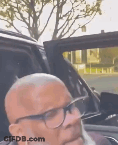 A Man With Glasses And A Beard Is Sitting In The Back Seat Of A Car . GIF
