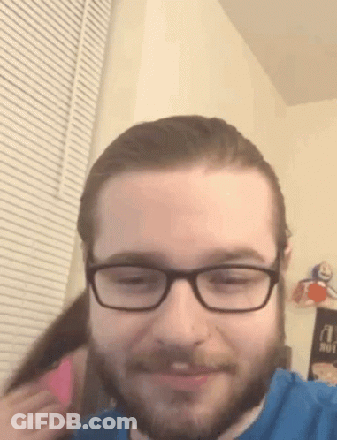 A Man With Glasses And A Beard Is Smiling While A Woman Holds His Hair . GIF