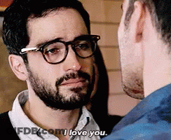 A Man With Glasses And A Beard Is Talking To Another Man Who Is Wearing A Blue Shirt . GIF