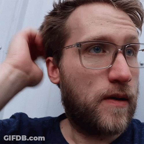 A Man With Glasses And A Beard Looks At The Camera With His Hand In His Hair GIF