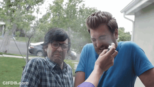 A Man With Glasses And A Blue Shirt Looks At Another Man With A Cigarette In His Mouth GIF