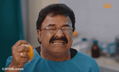 A Man With Glasses And A Mustache Is Holding An Orange In Front Of A Sign That Says Xmk GIF