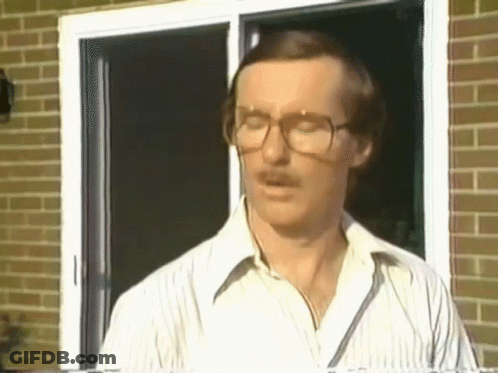 A Man With Glasses And A Mustache Is Standing In Front Of A Brick Wall . GIF