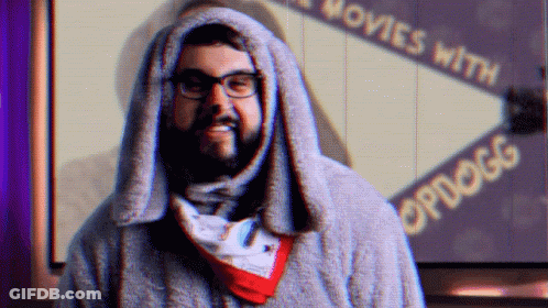 A Man With Glasses And A Scarf Around His Neck Stands In Front Of A Sign That Says Movies With Opdogg GIF