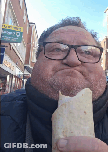 A Man With Glasses Is Eating A Burrito In Front Of A Sign That Says ' Shop Ot 9Oh ' On It GIF