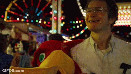 A Man With Glasses Is Holding A Stuffed Bird At A Carnival GIF