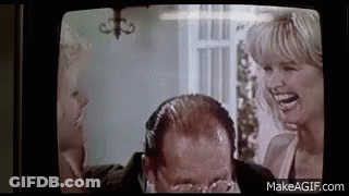 A Man With Glasses Is Laughing While A Woman Laughs Behind Him On A Television Screen . GIF