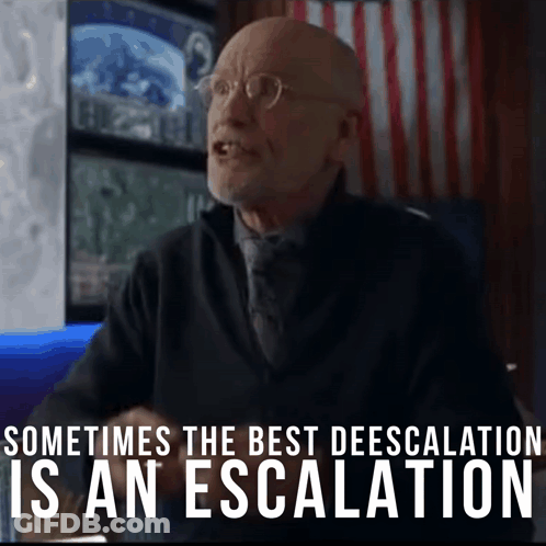 A Man With Glasses Is Sitting In Front Of A Sign That Says Sometimes The Best Deescalation Is An Escalation GIF