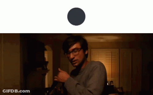 A Man With Glasses Is Standing In A Dark Room With A Black Circle Behind Him GIF