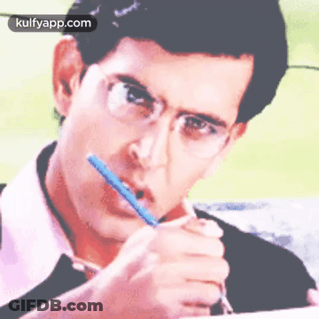 A Man With Glasses Is Writing On A Piece Of Paper With A Pencil In His Mouth . GIF