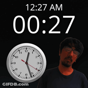 A Man With Glasses Stands In Front Of A Clock That Says 12:27 Am GIF