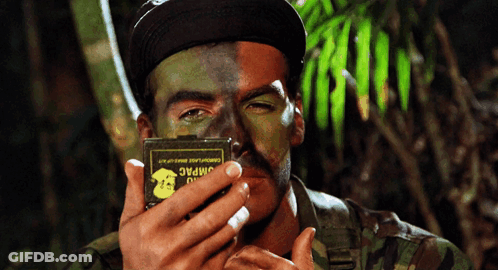 A Man With Half His Face Painted In Camouflage Holds A Box That Says Jimpac GIF