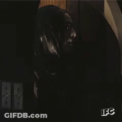 A Man With Long Black Hair And Glasses Is Standing In A Dark Room With A Sign That Says Ifc GIF