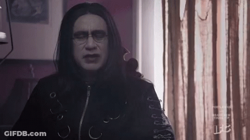A Man With Long Black Hair And Glasses Is Standing In Front Of A Window In A Room . GIF
