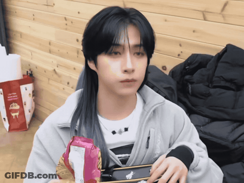 A Man With Long Black Hair Is Holding A Bag Of Chips In Front Of A Bag That Says Crayon Shin Chan GIF