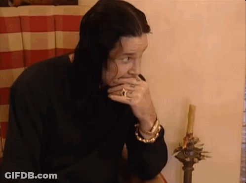A Man With Long Black Hair Is Sitting At A Table With His Hand To His Mouth . GIF