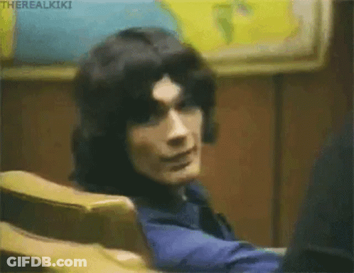 A Man With Long Black Hair Is Sitting In A Chair And Looking At The Camera . GIF