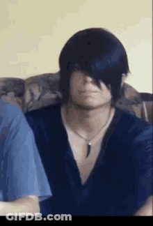 A Man With Long Black Hair Is Sitting On A Couch Wearing A Blue Shirt And A Necklace . GIF