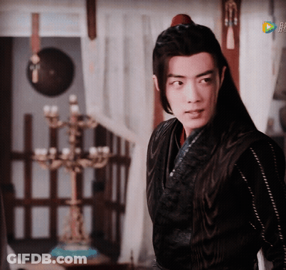 A Man With Long Black Hair Is Standing In A Room With A Candelabra In The Background GIF
